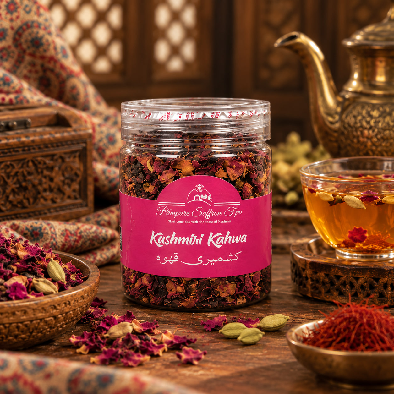 A jar of Kashmiri Kahwa with dried rose petals and spices, a brass kettle behind it and a glass of brewed kahwa beside it, bowls of cardamom and saffron in front