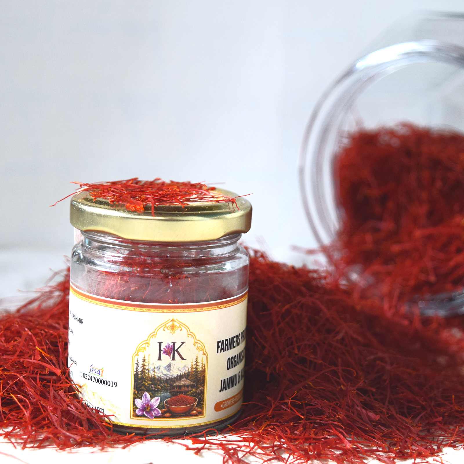 The Heritage Kashmir glass jar of Kashmiri Mongra saffron on a heap of red threads, a tipped glass jar spilling more saffron behind it