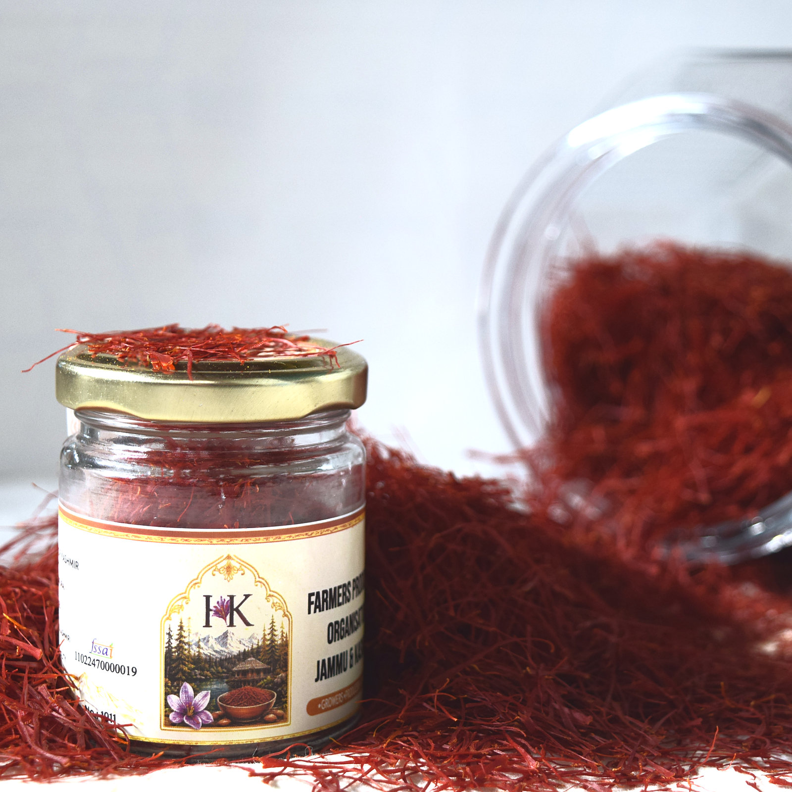 The saffron jar with its Farmers Producers Organisation Jammu & Kashmir label, beside a tipped glass jar pouring deep-red Mongra threads onto the table