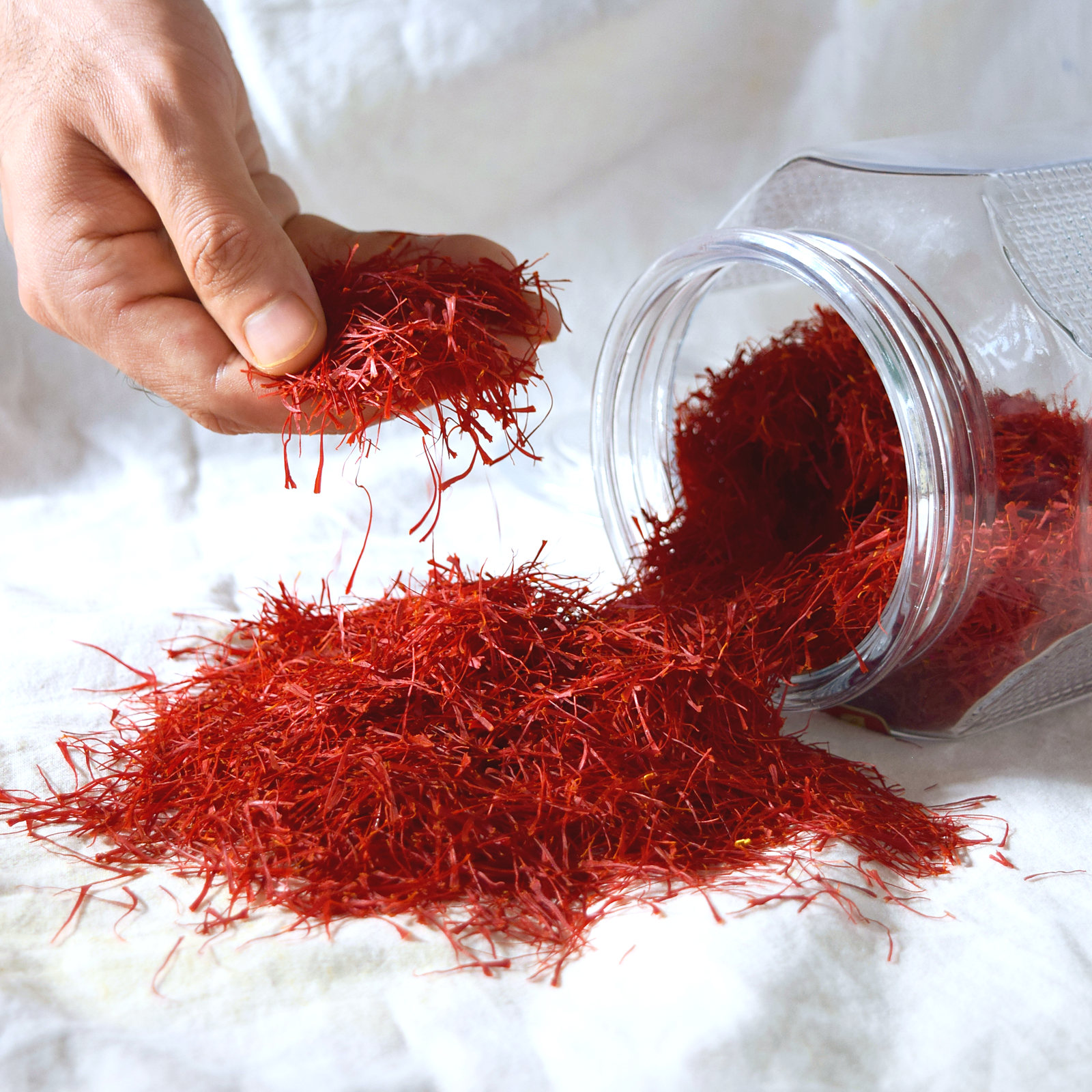 A hand letting long red Kashmiri saffron threads fall onto a heap spilling from a glass jar on white cloth