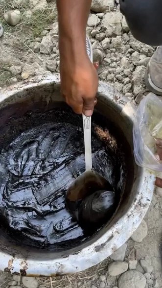 A hand stirring dark shilajit resin in a metal bowl set over a fire