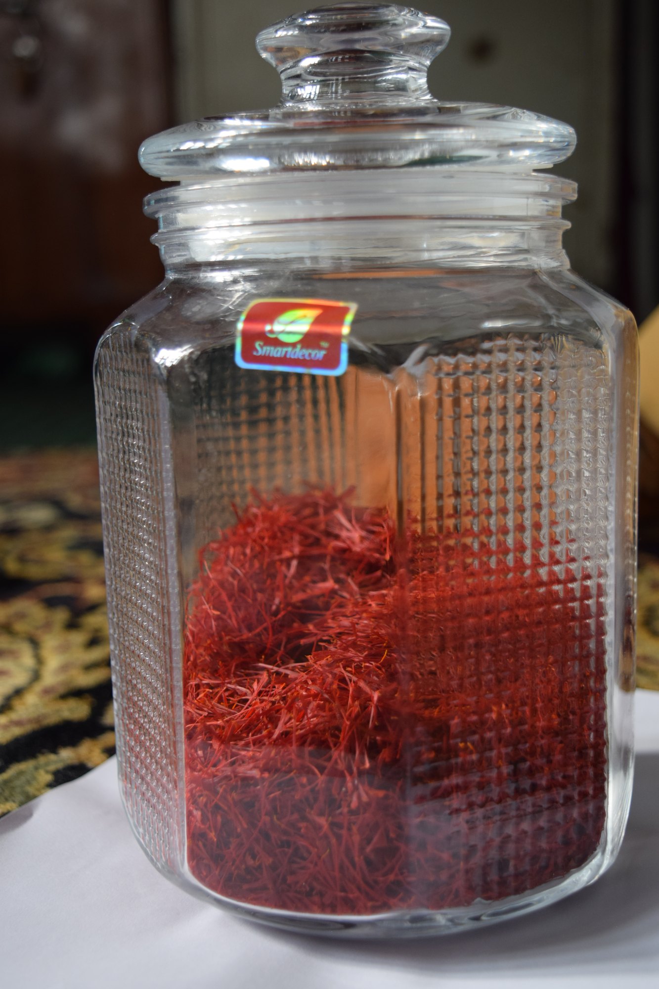 A tall ribbed glass jar filled with loose red saffron threads, standing on a patterned cloth